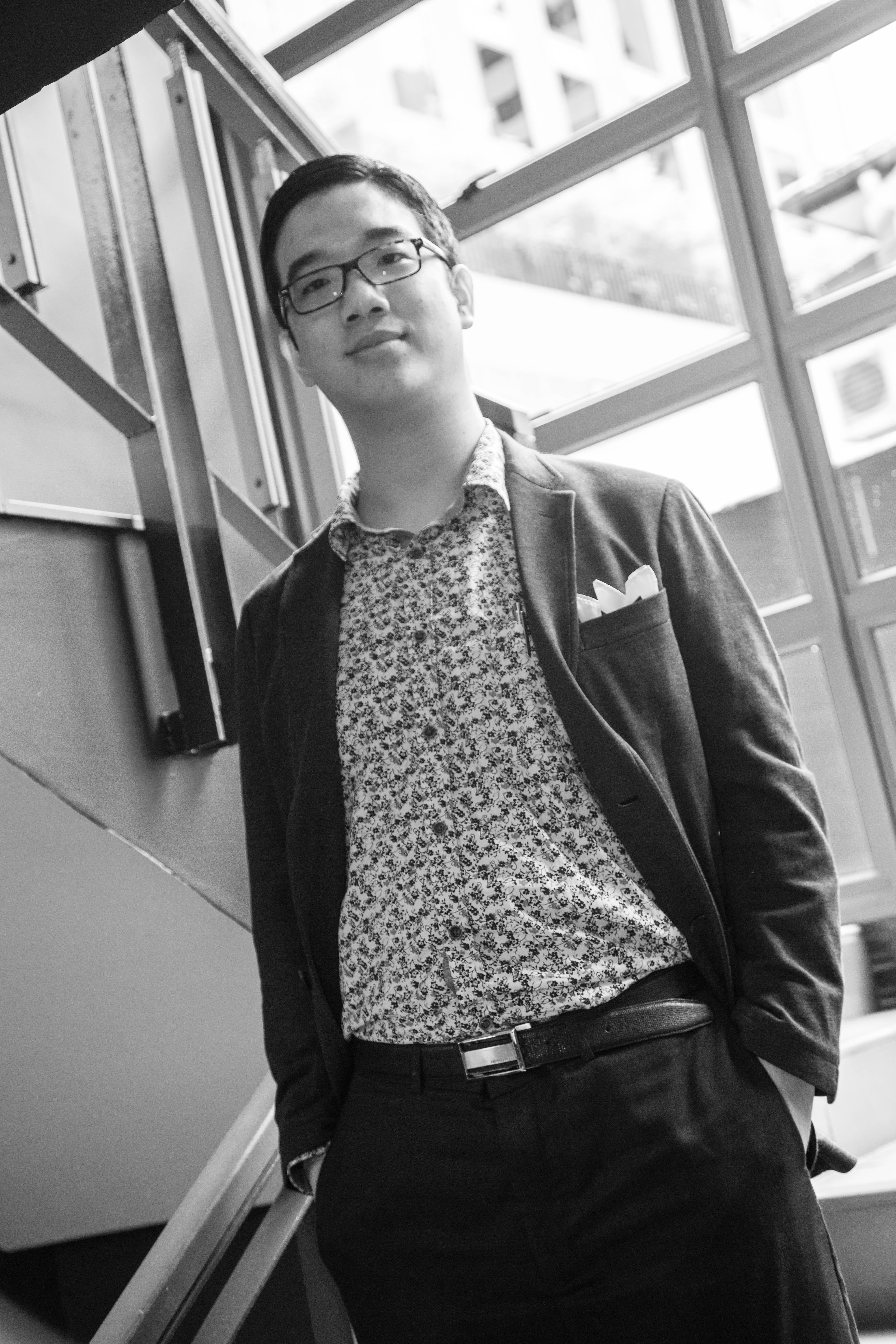 A male-presenting person wearing glasses, in a patterned shirt and black jacket, standing on some stairs.