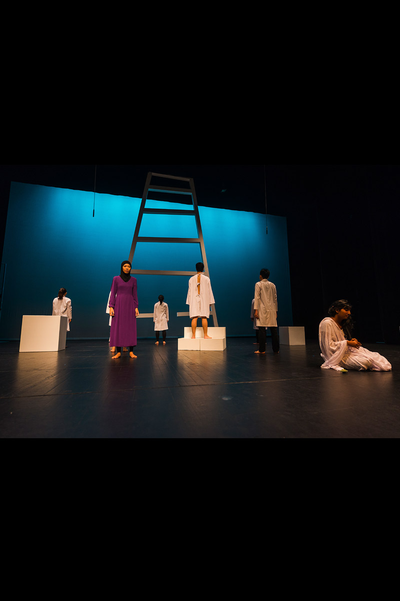 Production Still of Furthest North Deepest South (2015): 7 performers positioned apart from one another on stage, 5 face upstage and 2 face downstage. On the left 1 female performer stands, dressed in a long purple dress and a black tudung. On the right a long-haired performer sits on the floor dressed in a long white dress. A giant ladder is in the centre as part of the set.