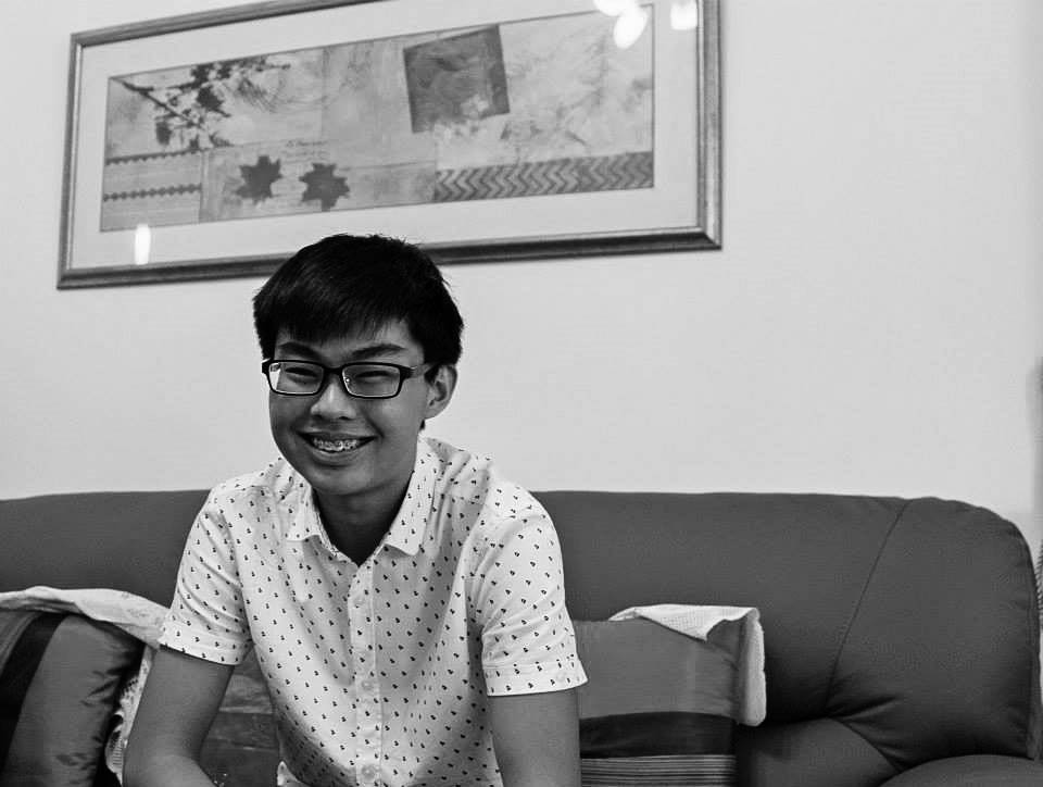 A male-presenting person wearing glasses and a white shirt with black dots. He is seated on a couch against a white wall with a painting above him.