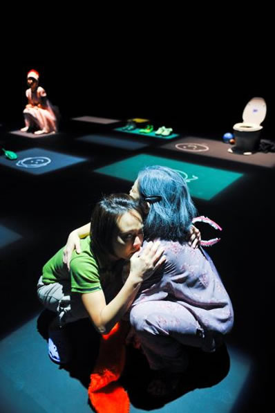 Production Still of Poop! (2010): A scene on stage lit with green and blue squares of light on the stage floor. A pair of actors, a younger girl and an older granny, squat in the foreground, in a tight embrace. A lone toilet bowl is in the background on the right.