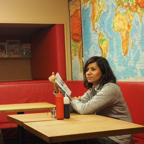 A female-presenting person seated on a long red seat, elbows on a yellow table. Behind her is a map of the world. She is holding a book in her right hand and looking out into the distance.