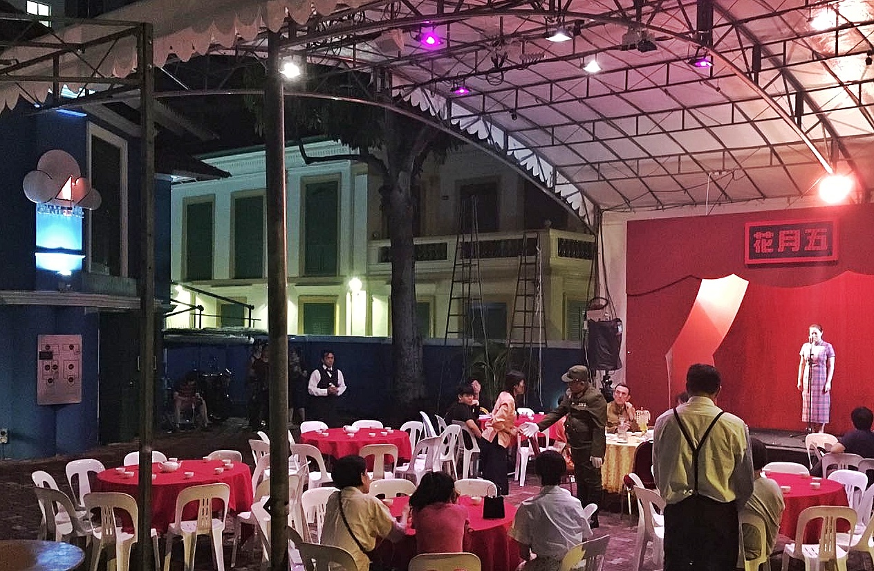 The front courtyard of 42 Waterloo Street with a white tentage erected. There are a number of tables dressed with red cloth and ports of tea and cups, with some audience members sitting at them watching a lady on a built stage.