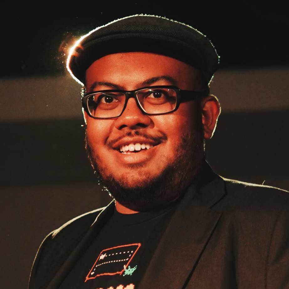 A smiling male-presenting person wearing a flat black cap, black glasses, a black shirt and black jacket.