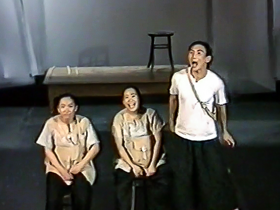 A scene from "Three Children": two performers sit on stools beside a third dressed in a white top and black pants, standing up with his mouth open.