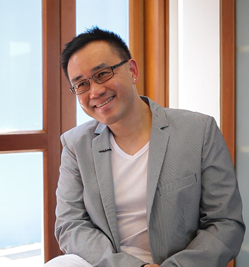 A short-haired man in a grey suit and white shirt, wearing glasses. His head is slightly tilted to the left and he is smiling.