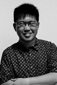 A black and white image of a bespectacled guy, wearing a short sleeved buttoned up shirt. He has his arms crossed and is smiling brightly.