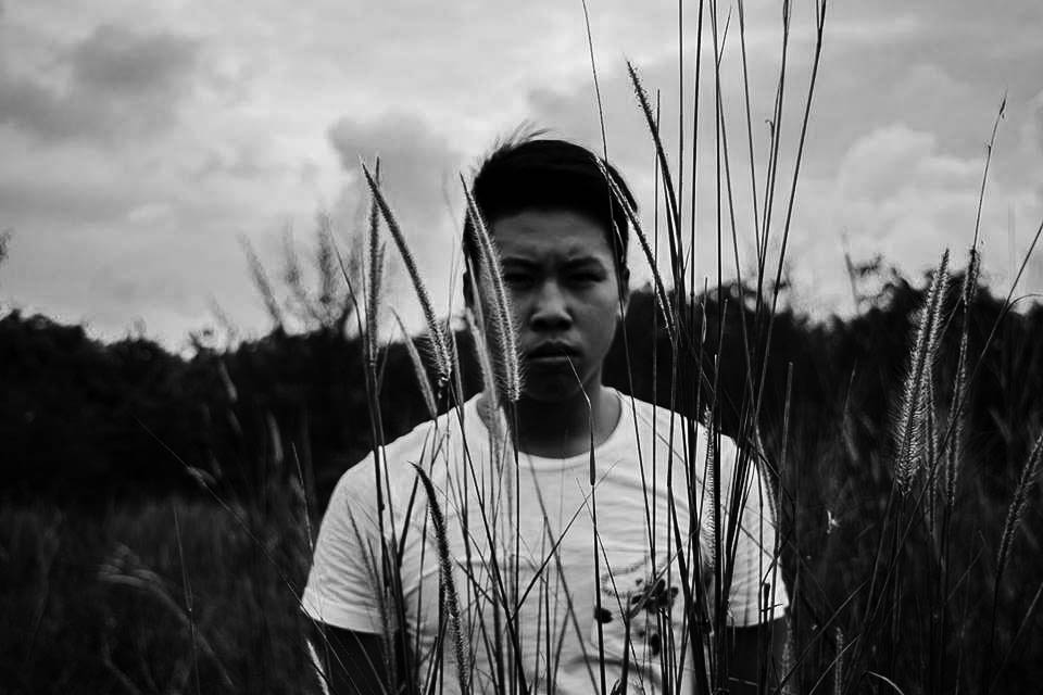 A black and white image of a guy frowning, while standing behind some leaves. 