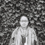 A black and white image of a bespectacled lady, glancing sideways downwards. She has her hair neatly tied up and is wearing a shawl.