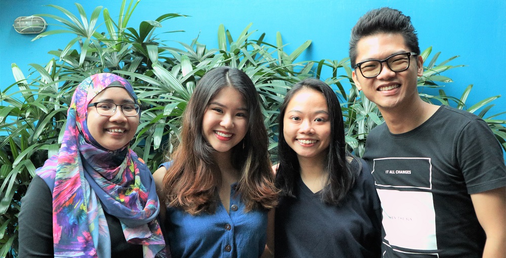 Four persons standing in front of a cluster of green plants in front of a blue wall. They are smiling into the camera.