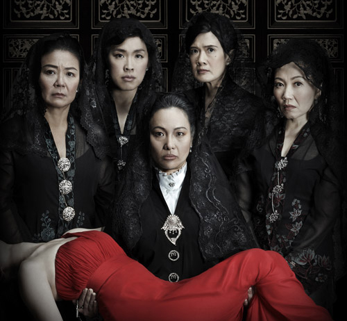 Set against a dark, patterned background with an ominous look, a female figure can be seen carrying the body of a woman donned in a dark red dress, while four female figures stand behind her. All of them carry a straight, stern expression. 