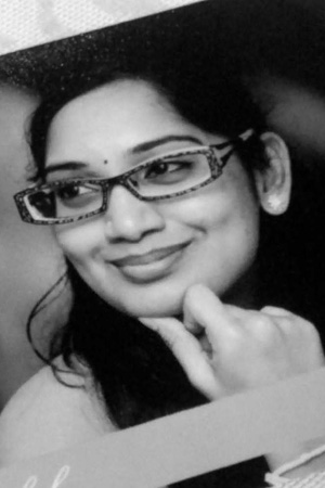 A black and white image of a bespectacled lady. She is smiling while looking to the side, and has her fingers on her chin.