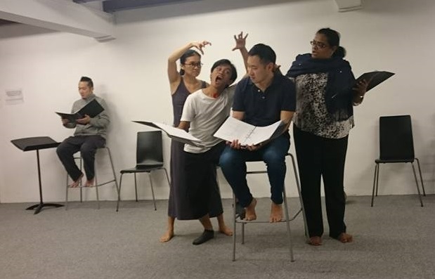 The photo is taken in Centre 42's rehearsal studio, and there is a person holding an open file seated on a high chair. Three people surround him, with two having one hand outstretched above their heads. There is another person seated in the corner on a high chair reading from a file.