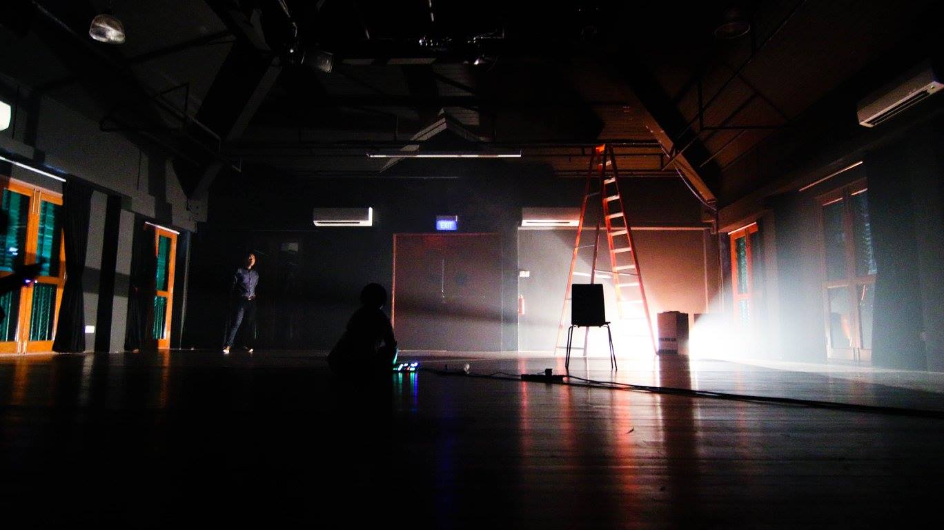 A black box space mostly dark, except for a light source on the ground lighting up two paths. A red ladder can be seen along with a chair in front that is backlit. To the left, an audience member dressed in blue can be seen.