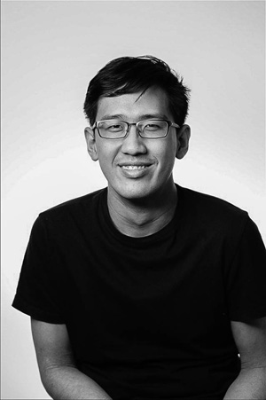 A black and white image of a bespectacled guy smiling, wearing glasses and a dark coloured shirt.