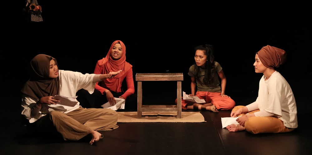 Four female-presenting persons seated on a stage, seated around a short wooden brown stool, each holding a piece of paper in their hands.