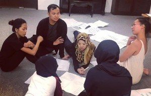 Six persons seated in a circle, speaking to one another, in a room with several pages of paper on the floor.