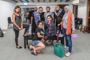 A group fo people standing around, and sitting on, a black bench in the 42 Waterloo Street Rehearsal Studio, smiling into the camera.