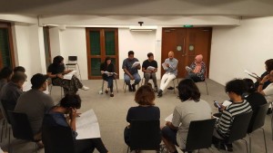 A group of people seated in a semicircle in the Centre 42 Rehearsal Studio, listening to five seated performers in front of them.