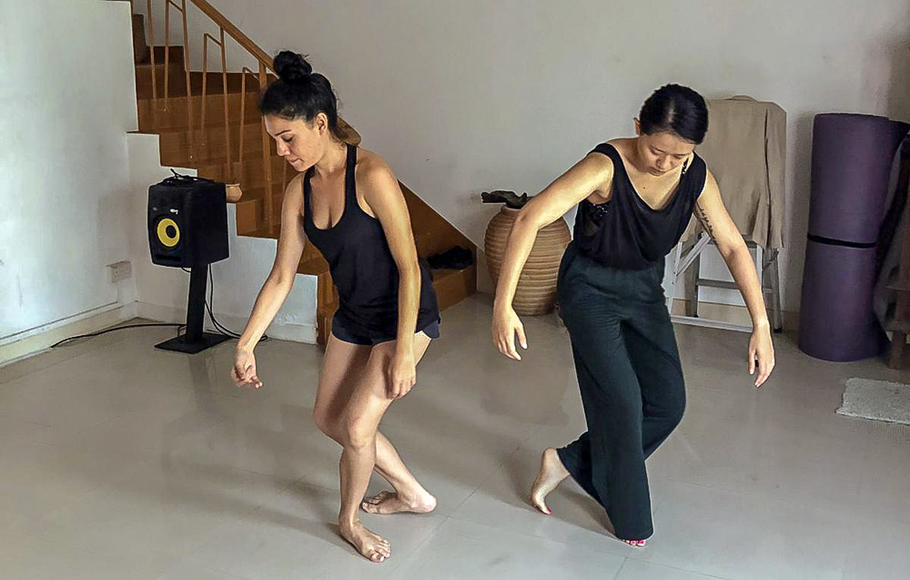 Two female-presenting persons in black clothing, practicing dance movements in a house.