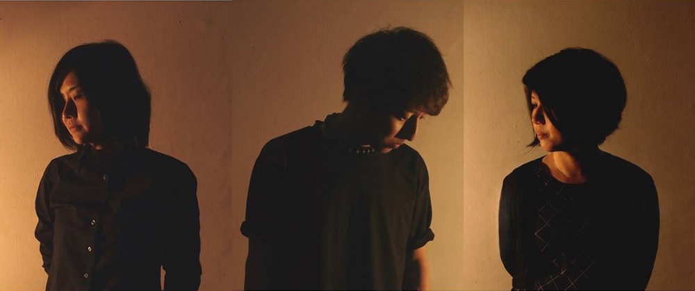 Three persons in black shirts standing against a brown wall, heads tilted downward and looking away from the camera.