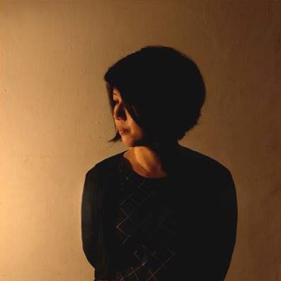 Against a yellow background, a female with short hair glances to the side, dressed in a black shirt.