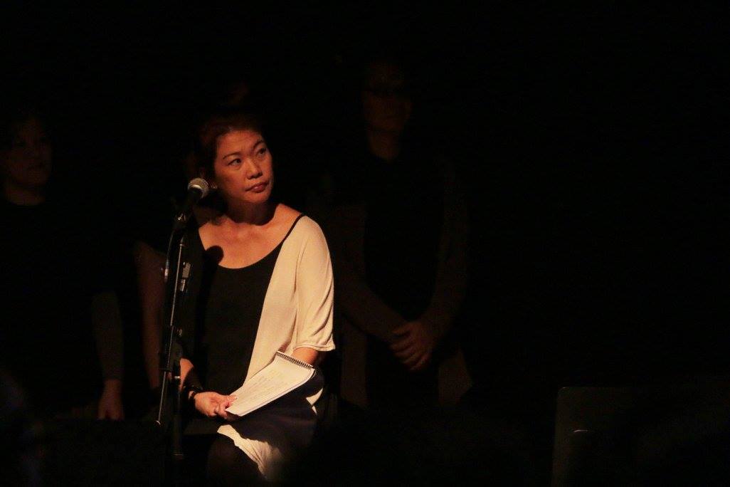 Photograph of a woman seated in a dimly lit black box, for The Vault: Leng-Geh-Mng 龍牙門.