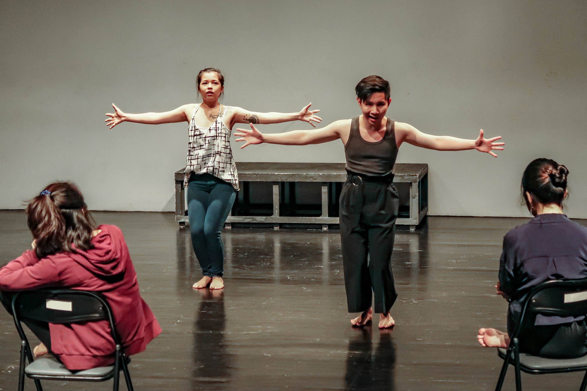 Two persons in sleeveless tops and pants stand barefoot on a stage, with their arms outstretched. In front of them are two people seated in chairs watching them.