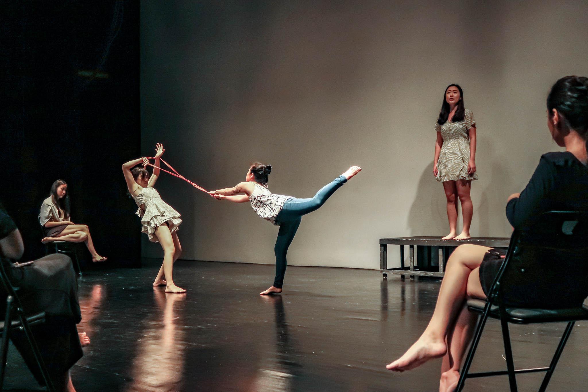 Two persons standing barefoot on a stage; one stands on on leg with the other lifted, hanging on to a long pink ribbon, while the other stands a distance in front of her with the ribbon around her wrists, leaning back and pulling away. Behind them is another person standing on a brown structure, looking out and speaking.