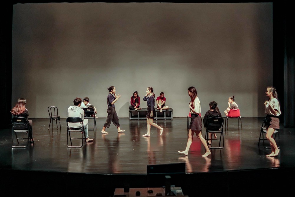 A group of performers on a stage. Several are seated in chairs around the stage, with two seated on a box at the rear of the stage. Four performers walk across the stage, one headed right and three headed left.