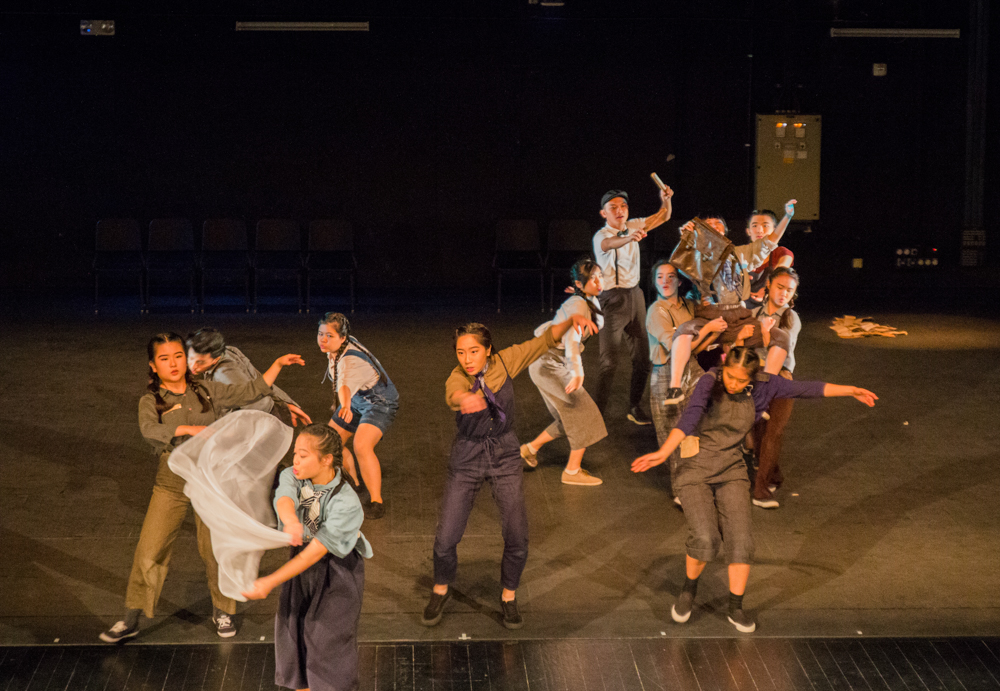 A group of people, mostly in brown and purple shirts and pants, gathered on a stage. To the right, four people hoist another in their arms, and in front of them are several more flailing their arms.