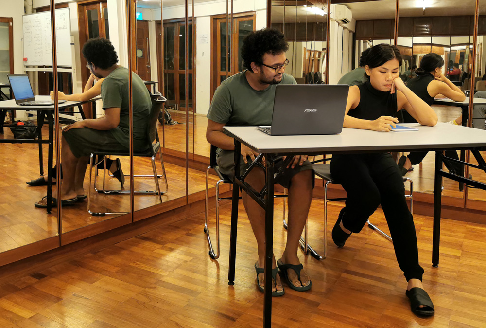 Two persons, one male-presenting and one female-presenting, seated side by side at a table in a corner of the 42 Waterloo Street Meeting Room, in front of several mirrors. There is a laptop on the table.