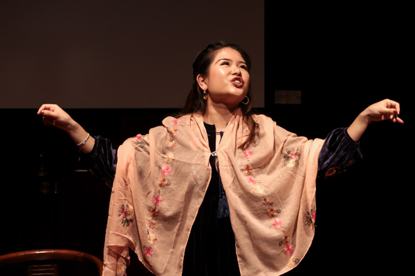 A female-presenting person draped in a light pink shawl with a pink flower pattern, with both her arms out to her sides, facing upwards.