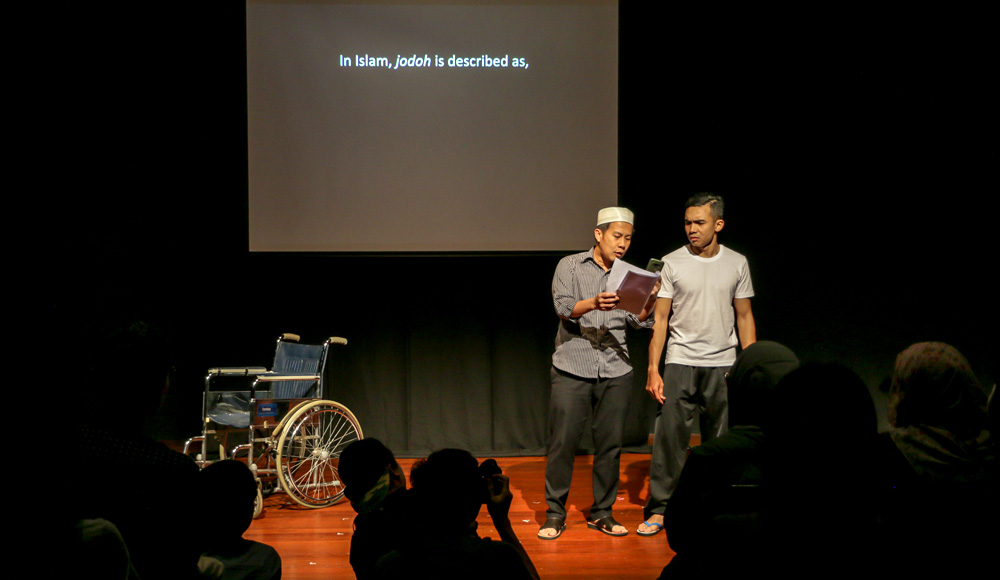 Two male-presenting persons stand on a stage in slippers. The person on the left holds out a sheaf of paper and shows it to the person on the right. To the left on the stage is a wheelchair. There is a screen behind displaying the words 'In Islam, jodoh is described as,'.