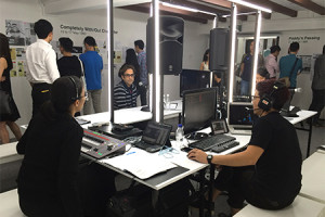 Several people milling around a room with exhibition panels on the wall. In the middle are tables set up in a rectangular fashion, with two persons in black clothing and headphones seated in front of laptops and other electronic equipment on the tables.