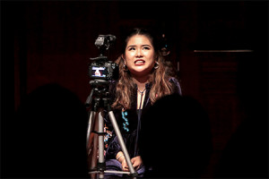 A female-presenting person seated in front of a camera on a tripod, speaking while glancing up to the left.