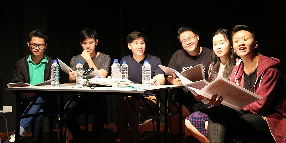 A row of six people seated at a table covered in paper and water bottles, reading from their scripts.