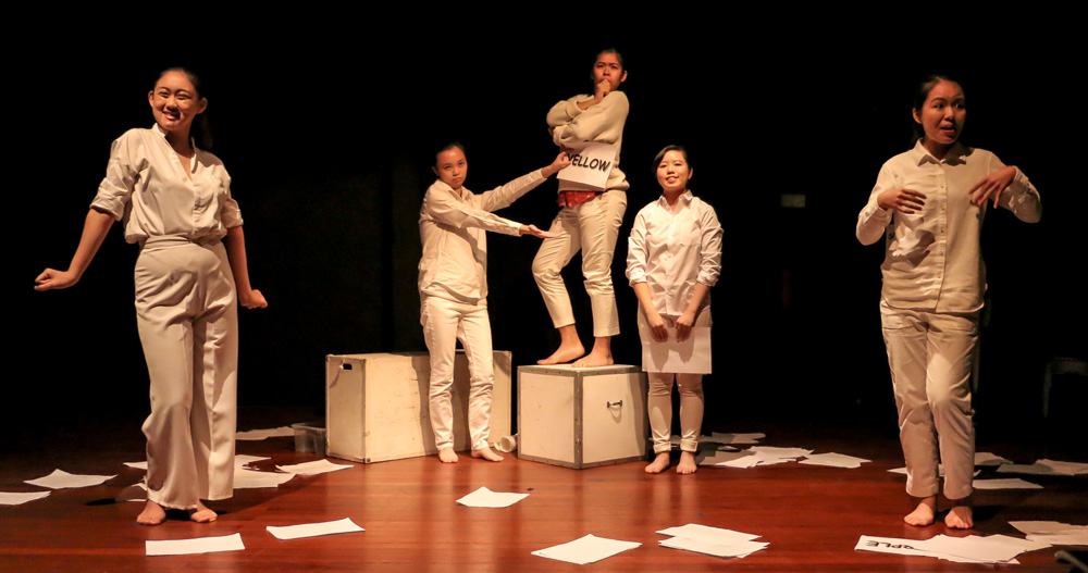 Five persons in white clothing, standing on a stage littered with white paper. Two stand in the foreground. In the background are two more, standing in front of some white boxes, while the last person stands on top of a white box with their arms folded.