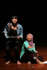 Two female-presenting persons, one wearing a denim jacket, half-kneeling and staring forward, the other on her right wearing a long-sleeved green top, seated cross-legged and looking up at her.