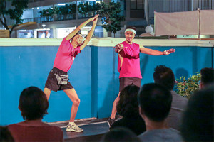 Two persons in bright pink exercise tops performing in the front courtyard at 42 Waterloo Street. The person on the left stretches out both arms towards the sky, palms pressed together; the person on the right gestures towards the audience.