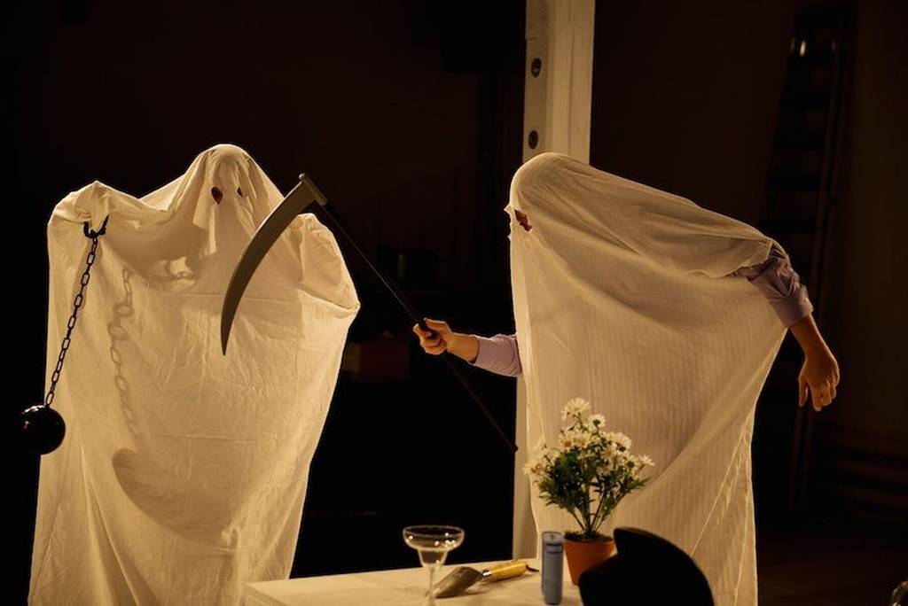 Two persons draped in white sheets to resemble ghosts. The one on the left holds a ball and chain. The one on the right wields a scythe. In the foreground is a table, on which are flowers and an empty margarita glass.