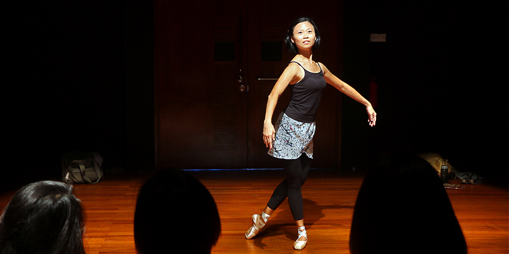 Jocelyn Chng in a sleeveless black top and ballet shoes, dancing in the 42 Waterloo Street Black Box in front of an audience.