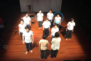 An aerial shot of a group of persons in white shirts and black pants, standing on a lit stage. One person towards the right is circled in blue.