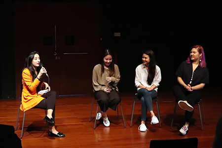 Four persons seated in a row in the 42 Waterloo Street Black Box. The person on the far left is in yellow, holding a microphone and speaking to the audience.