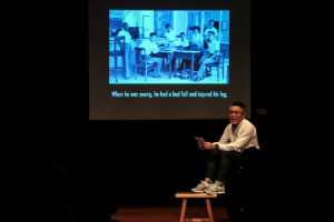 A male-presenting person in a long-sleeved white jacket seated on a black box with their feet on a brown stool. Behind them is a blue-tinted image projected on a projector screen.