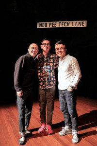 Three male-presenting persons standing in a row, arm around each other and smiling. Behind them against a black wall is a sign with the words 'NEO PEE TECK LANE'.