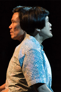 Two persons seated side by side, facing opposite directions. The person in front is in a patterned blue shirt, facing right while speaking. Behind them is another person, facing left with a neutral expression.