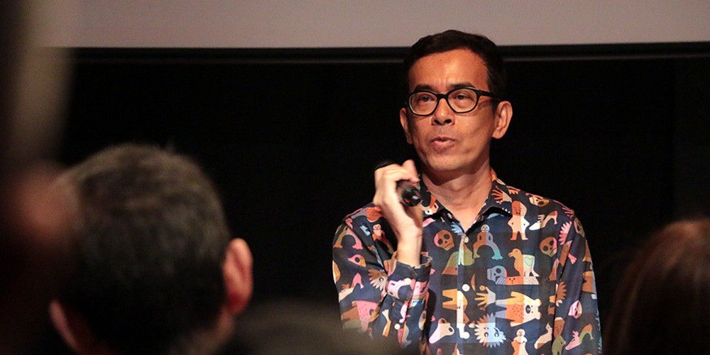 Neo Kim Seng in a colourful top, holding a microphone, speaking to an audience.