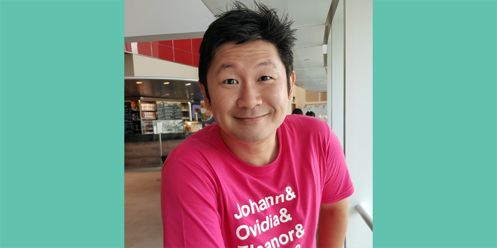 Ng Yi-Sheng in a pink shirt, smiling into the camera. The photograph is bordered by green rectangles on either side.