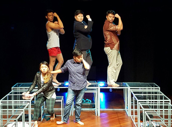 Several glass tables set up in a reverse U-shape on a stage. Three masculine-presenting persons stand on the bottom of the U with their right profiles facing the audience, right arms held up in a fist. In front of them on the stage are two more persons. The one on the left is seated on a glass table and the one on the right has their right hand on the other person's back.