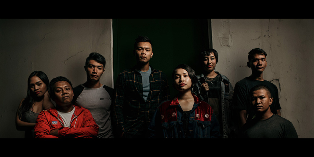 Eight people, mostly clad in jackets and t-shirts, standing in front of two grimy white walls, staring into the camera.